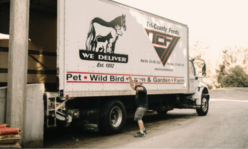 A Tri-County Feeds branded box truck with a staff member posed in front of it on a clear day.